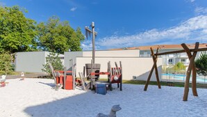 A sandy outdoor area with a swing set, a wooden structure, and a pool in the background. - Instant Evasion - Piscine Partagée (La Teste-de-Buch)