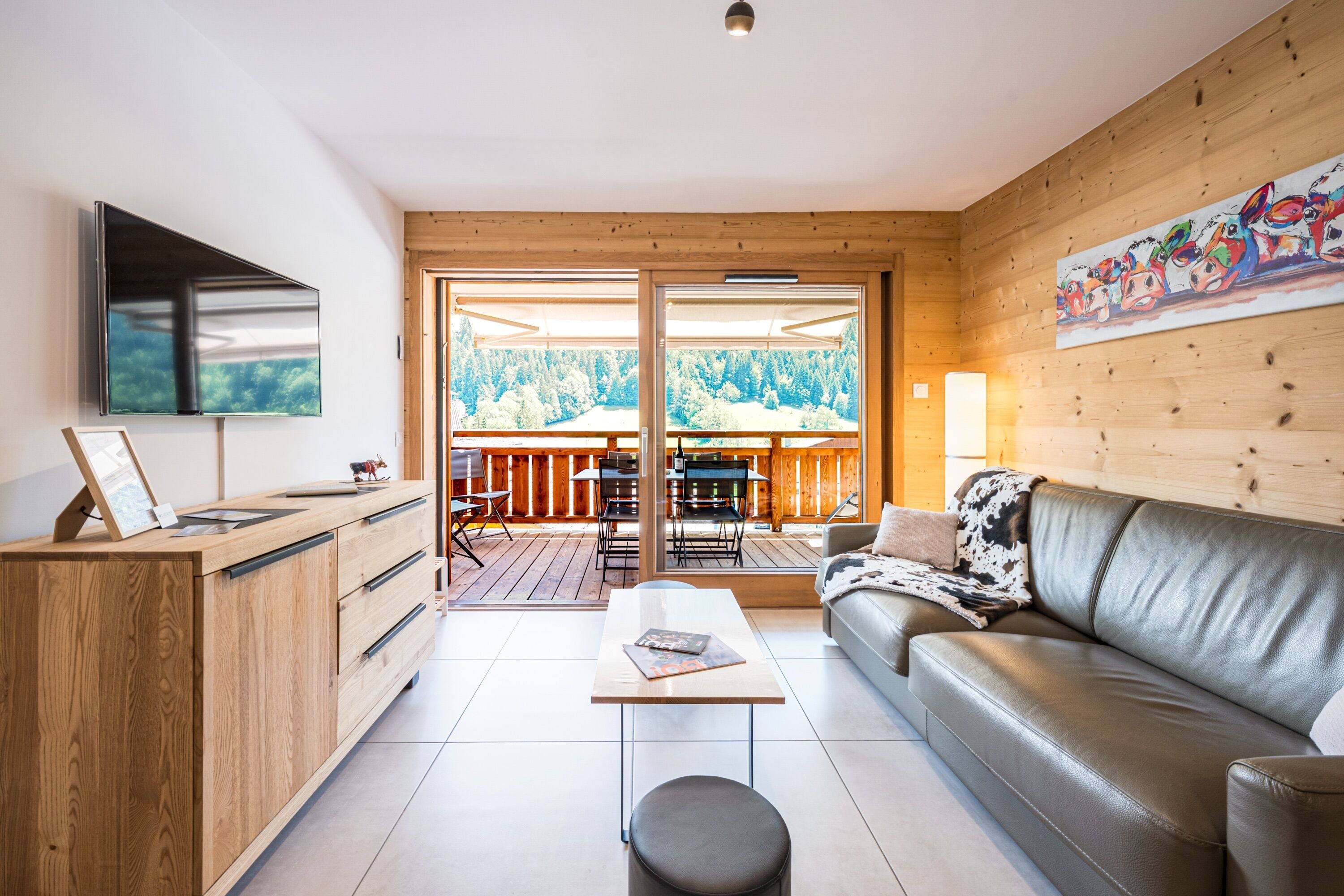Ein modernes Wohnzimmer mit Holzboden, einer Ledersofa, einem Flachbildfernseher, einem Essbereich mit Outdoor-Möbeln und einem großen Fenster mit Blick auf einen Wald.