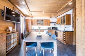 A modern kitchen with wooden cabinets, a dining table with blue chairs, a built-in TV, and a large window. - Mont Blanc - Apt N°1 (Le Grand Bornand)