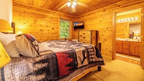 A wooden cabin bedroom with a bed, a ceiling fan, a television, and a bathroom with a vanity. - Full Arcade Game Room, Hot Tub & Mountain Views, Sleeps 10 Near Dollywood (Sevierville)