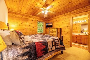 A wooden cabin bedroom with a bed, a ceiling fan, a television, and a bathroom with a vanity. - Full Arcade Game Room, Hot Tub & Mountain Views, Sleeps 10 Near Dollywood (Sevierville)