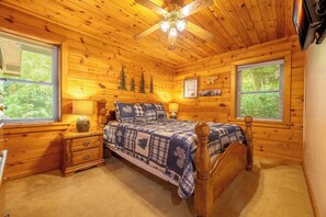 A wooden cabin bedroom with a bed, nightstands, a ceiling fan, and windows. - Full Arcade Game Room, Hot Tub & Mountain Views, Sleeps 10 Near Dollywood (Sevierville)