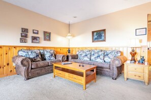 A living room with a wooden paneled wall, a brown leather sofa with patterned cushions, a wooden coffee table, and a wooden nightstand with a lamp. - Full Arcade Game Room, Hot Tub & Mountain Views, Sleeps 10 Near Dollywood (Sevierville)