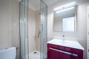 A bathroom with a glass-enclosed shower, a white toilet, a red vanity with a white sink, and a mirror above the sink. - Le Cocon du Barcarès - Proche Plage (Le Barcarès)