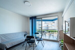 A modern living room with a sofa, a glass table, and chairs, a TV, and a balcony with a view of other buildings. - Le Cocon du Barcarès - Proche Plage (Le Barcarès)