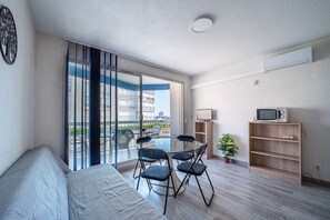 A modern living room with a glass door leading to a balcony, a sofa, a dining table with chairs, a TV, and a small plant. - Le Cocon du Barcarès - Proche Plage (Le Barcarès)