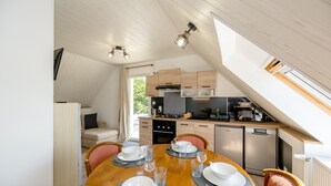 A dining area with a round table set for four, a kitchen with modern appliances, and a loft-style ceiling with skylights. - Au pas de côté plourhannais - Proche Plage (Plourhan)