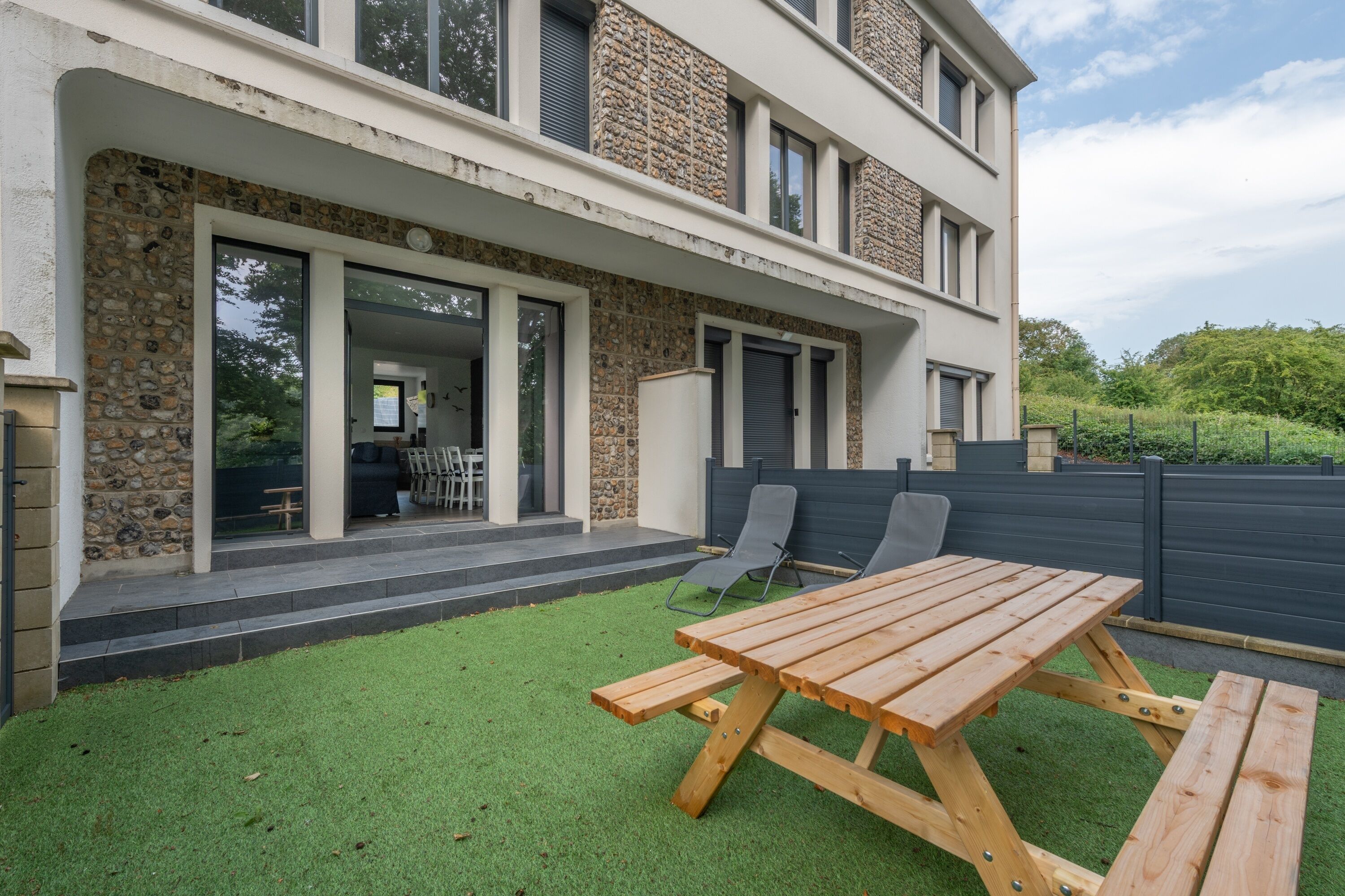 Een modern huis met een stenen gevel, een terras met een houten tafel en stoelen, en uitzicht op het omliggende groen.