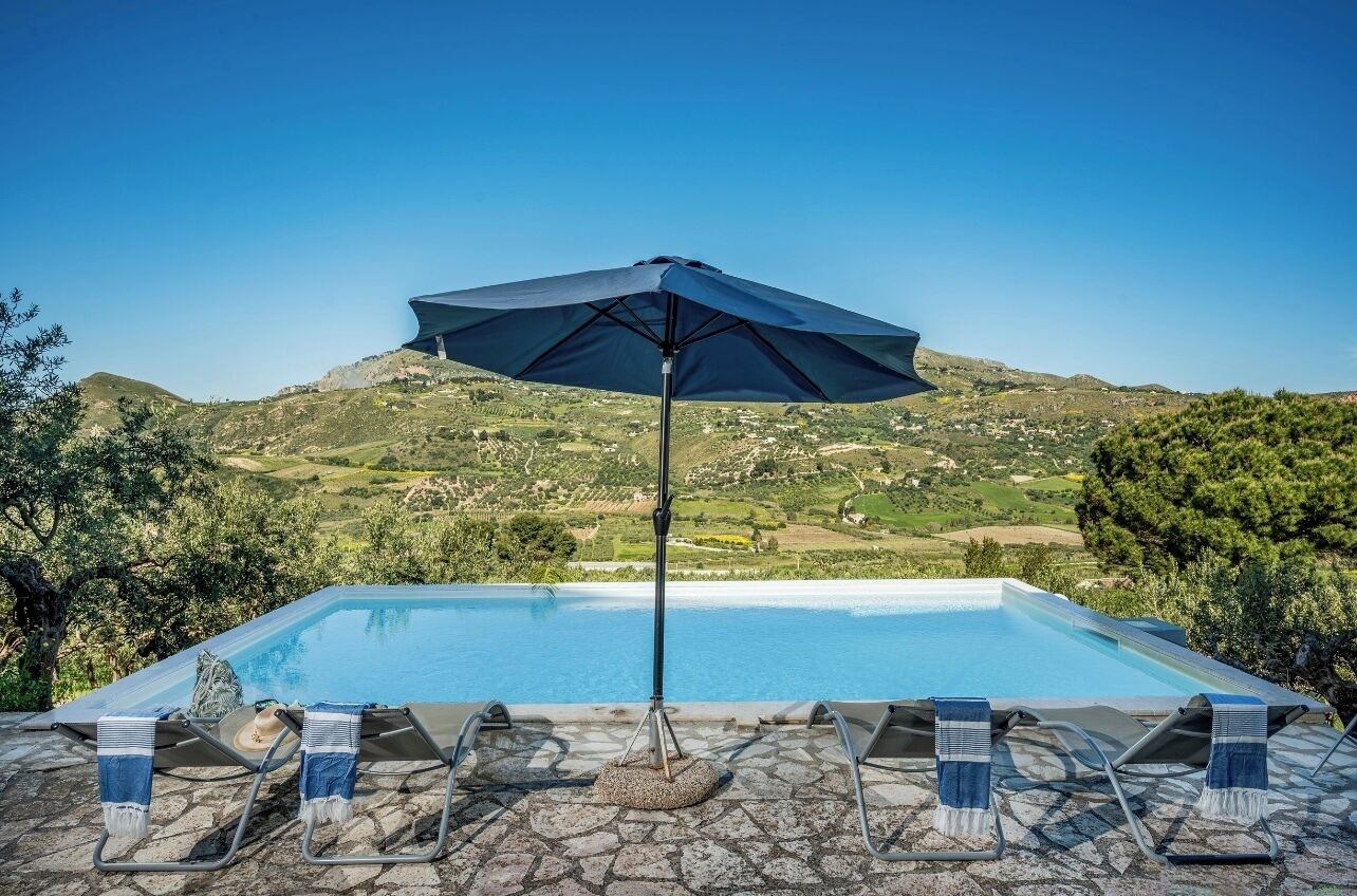 Une piscine entourée d’une terrasse en pierre, agrémentée de chaises longues et d’un parasol, et offrant une vue sur un paysage vallonné.
