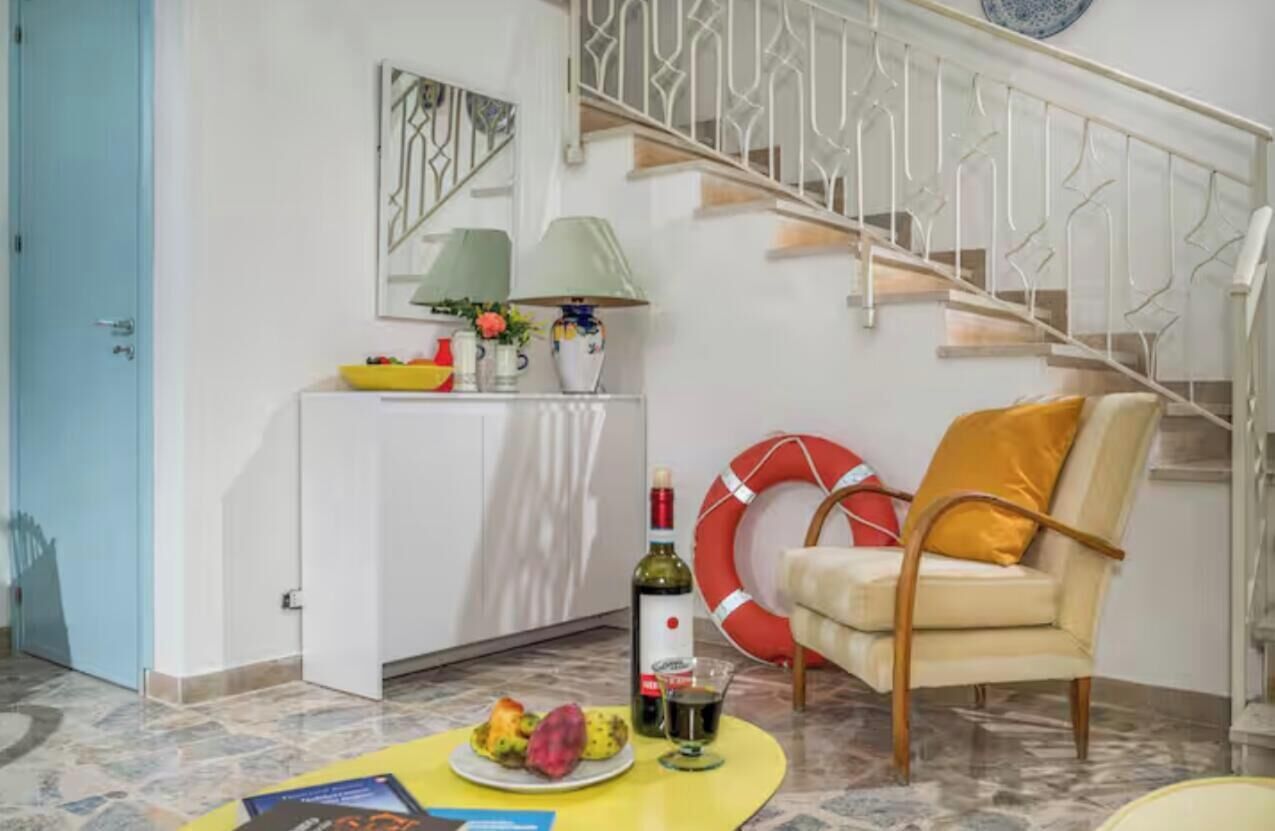 Ein Wohnzimmer mit einem weißen Sofa, einem gelben Tisch mit einer Fruchtschale, einer Weinflasche und einem roten Rettungsring auf dem Boden.