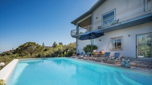 A swimming pool with lounge chairs and umbrellas, a balcony with a railing, and a building with large windows. - Villa Le Terrazze Blu (Sicilia)