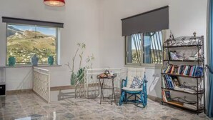 A room with a view of a mountainous landscape, a red pendant light, a blue chair, a small table, and a bookshelf. - Villa Le Terrazze Blu (Sicilia)