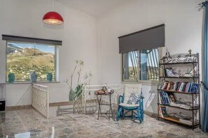 A room with a view of a mountainous landscape, a red pendant light, a blue chair, a small table, and a bookshelf. - Villa Le Terrazze Blu (Sicilia)