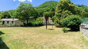 Un área residencial con dos casas, una de fachada amarilla y la otra de fachada blanca, rodeada de abundante vegetación y una colina al fondo.