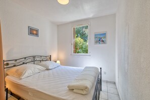 Ein Einzelbett mit weißer Bettwäsche in einem Zimmer mit weißen Wänden, ein Fenster mit Blick auf Grünfläche und zwei gerahmte Bilder.