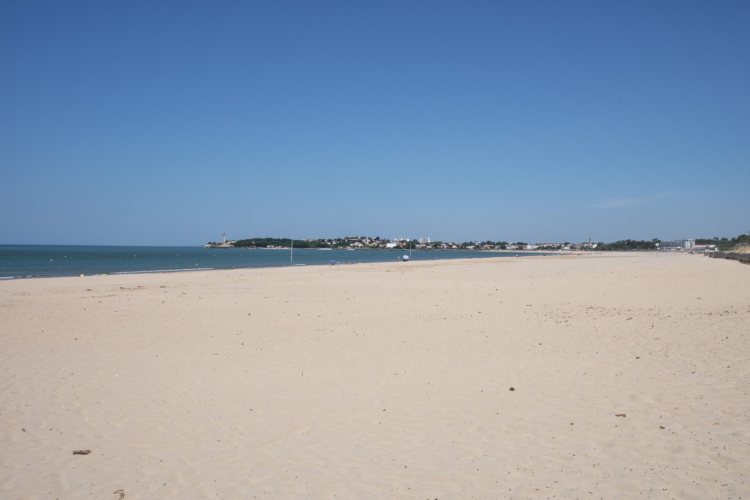 A wide sandy beach with a clear sky, a calm sea, and a distant shoreline with buildings.