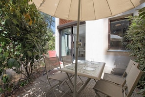 Een terras buiten met een tafel en stoelen, een glazen deur en uitzicht op de omliggende beplanting.