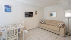 Ein Zimmer mit an der Wand montiertem Fernseher, einer Couch mit Kissen, einem Tisch mit Stühlen und einer Stehlampe.