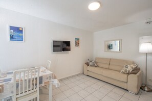 Ein Zimmer mit an der Wand montiertem Fernseher, einer Couch mit Kissen, einem Tisch mit Stühlen und einer Stehlampe.