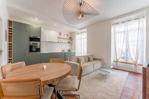 Eine moderne Küche mit integrierten Geräten, ein Essbereich mit rundem Tisch und Stühlen, eine Couch und ein großes Fenster mit Vorhängen.