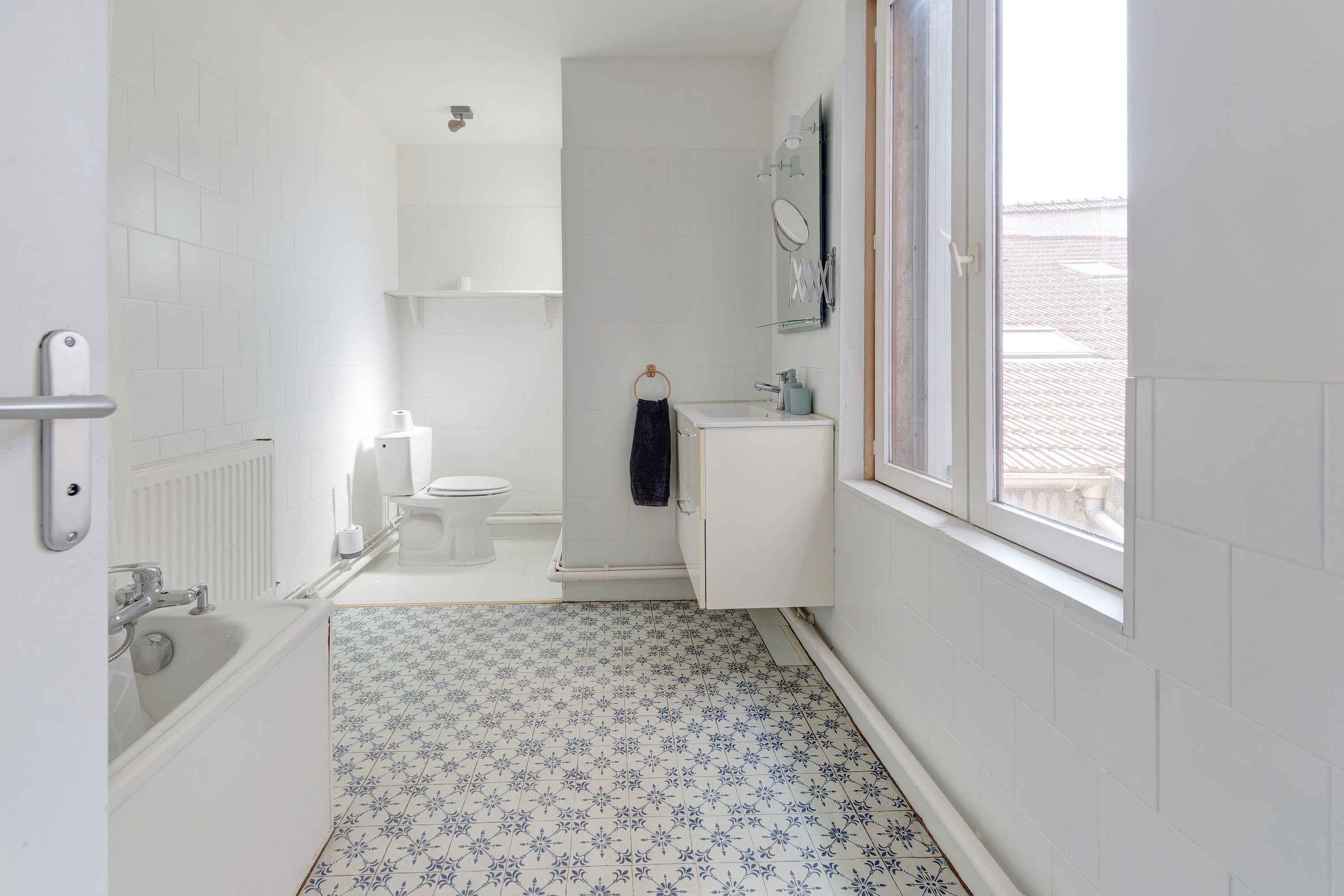 A narrow bathroom with white tiles, a bathtub, a toilet, and a window.