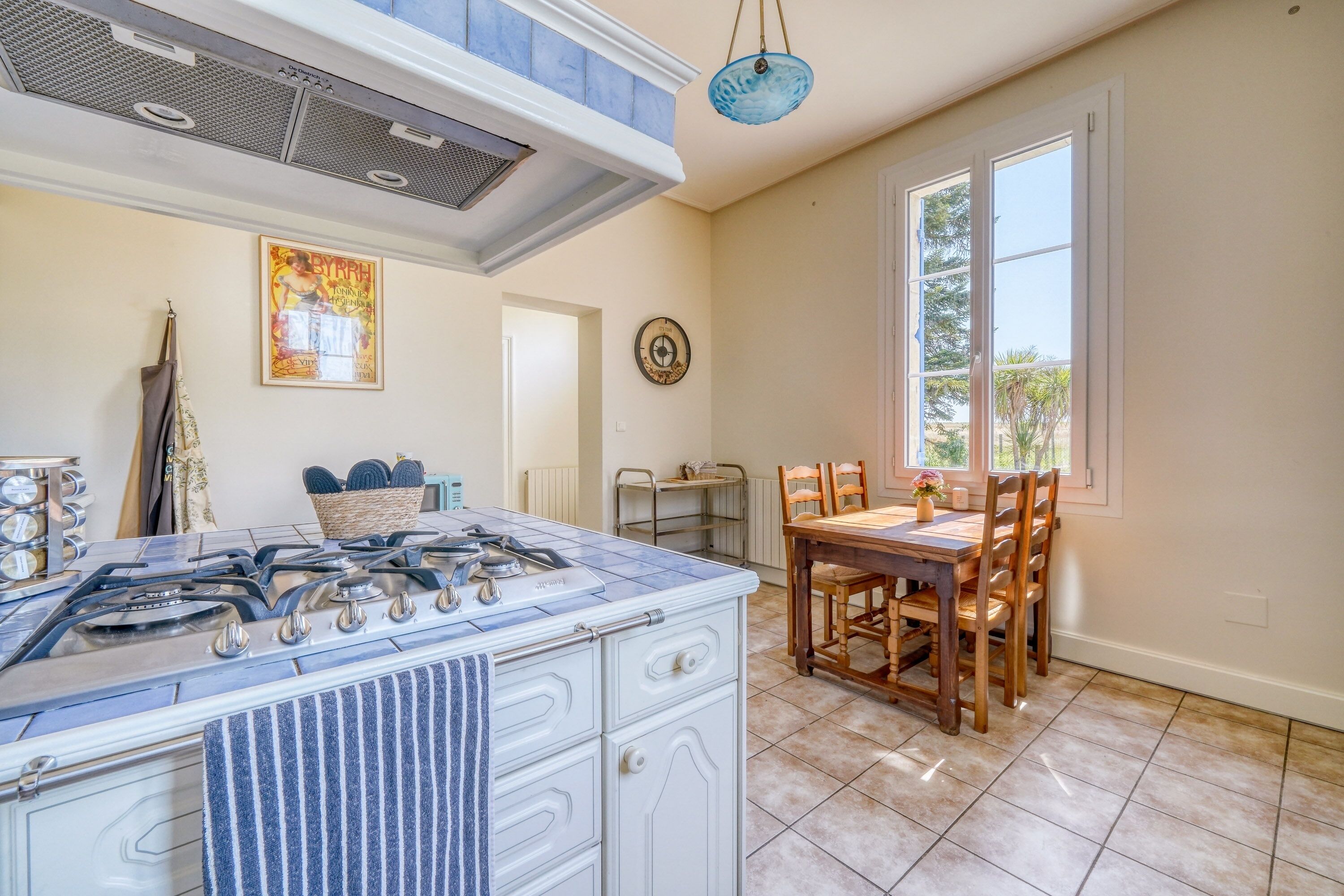 A kitchen with a gas stove, white cabinetry, a blue towel, a dining area with wooden chairs, and a window with a view of greenery.