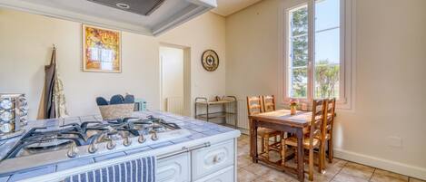 A kitchen with a gas stove, white cabinetry, a blue towel, a dining area with wooden chairs, and a window with a view of greenery.