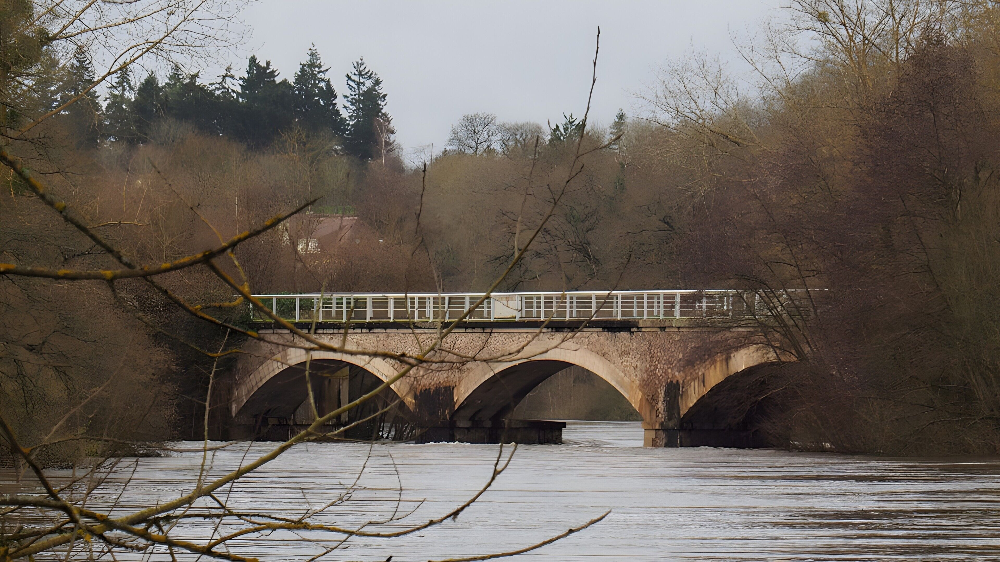 Un pont en pierre, composé de trois arches, enjambe un cours d’eau et est entouré d’arbres et de végétation.