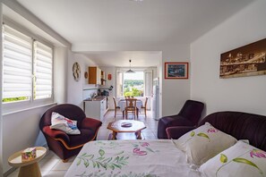 A room with a bed, two armchairs, a small table, a window with blinds, a kitchen area, and a dining table with chairs.
