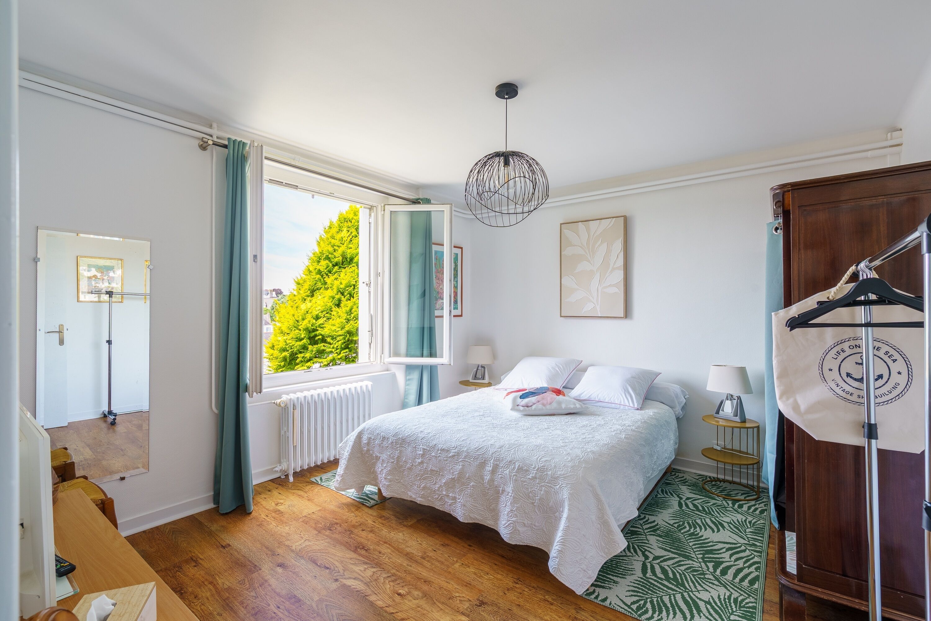 A bedroom with a bed, a window with curtains, a radiator, a hanging light fixture, a wardrobe, and a small bedside table with a lamp.
