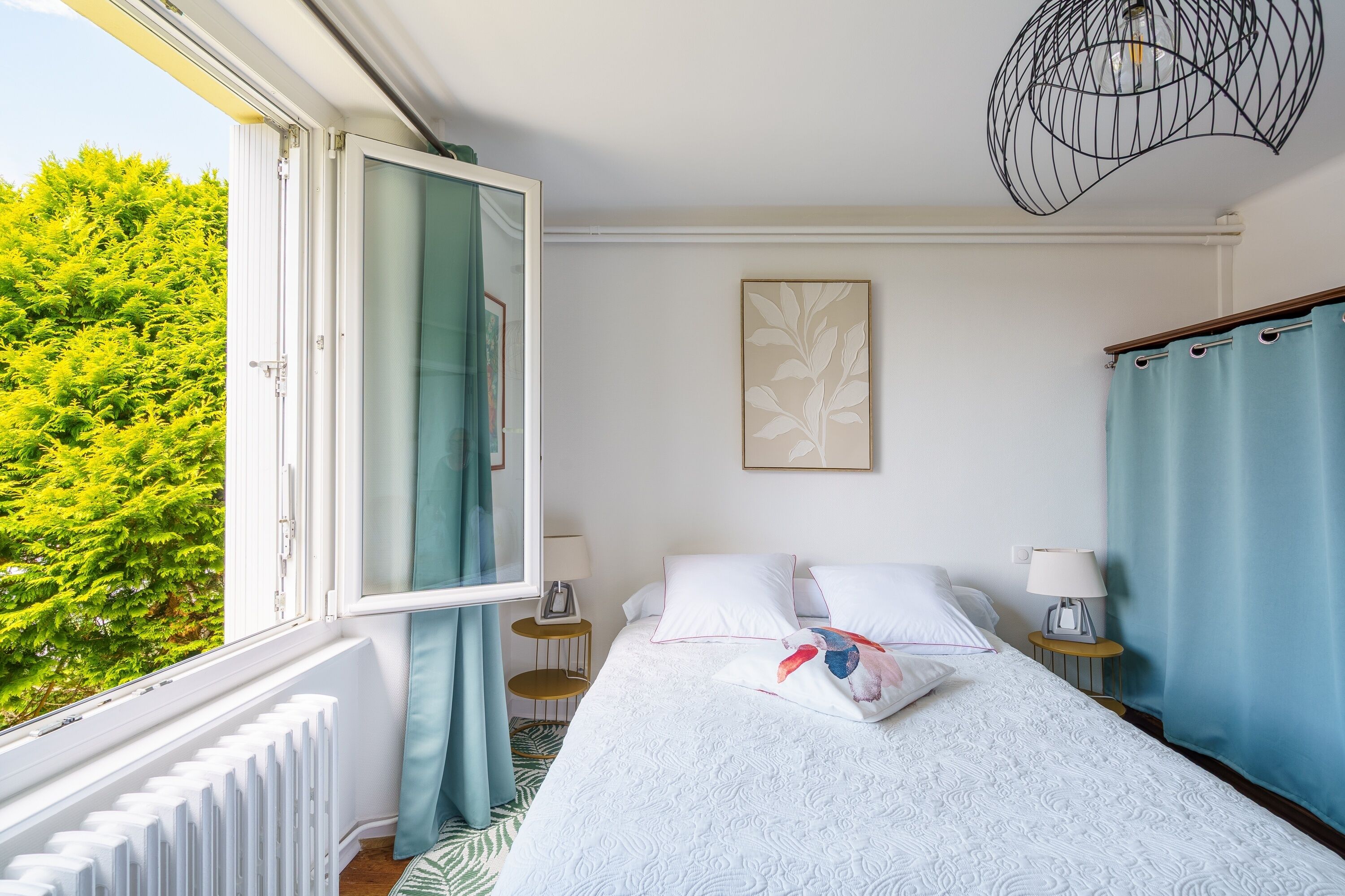 A hotel room with a bed, bedside tables, a window with curtains, a radiator, and a view of greenery outside.