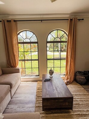 Ett vardagsrum med en soffa, ett soffbord i trä och två stora, välvda fönster med gardiner.