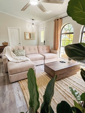 Ein Wohnzimmer mit einem beigefarbenen Ecksofa, einem Holztisch und großen Grünpflanzen.