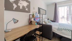 Ett välordnat sovrum med en säng, ett skrivbord med en lampa, en världskarta på väggen och ett fönster med gardiner.