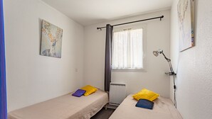 Pieni, siisti huone, jossa on kaksi erillistä sänkyä, ikkuna, jossa on ohuet verhot, patteri ja seinään kiinnitetty lamppu.