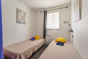Ein kleiner, sauberer Raum mit zwei Einzelbetten, einem Fenster mit durchsichtigen Vorhängen, einem Heizkörper und einer Wandlampe.