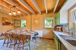 A dining area with a table set for four, wooden chairs, a painting on the wall, and a kitchen with a sink and a mixer. - Ty Crugo - Jolie maison familiale avec jardin (Saint-Lyphard)
