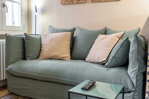 Ein grünes Sofa mit vielen Kissen, eine Fernbedienung auf einem kleinen Tisch und ein Fenster mit Jalousien.