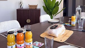 A dining table with various items including bottles of orange juice, a loaf of bread, and a cutting board. - Tranquiloue - Appart Centre-Ville 20min Puy du Fou (Les Herbiers)