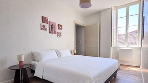 A bedroom with a bed, a small table, a red rug, a wall with pictures, a window, and a door. - Tranquiloue - Appart Centre-Ville 20min Puy du Fou (Les Herbiers)