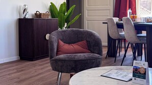 A modern living room with a round table, a sofa with a cushion, a dark cabinet, a potted plant, and a dining area with chairs and a table. - Tranquiloue - Appart Centre-Ville 20min Puy du Fou (Les Herbiers)