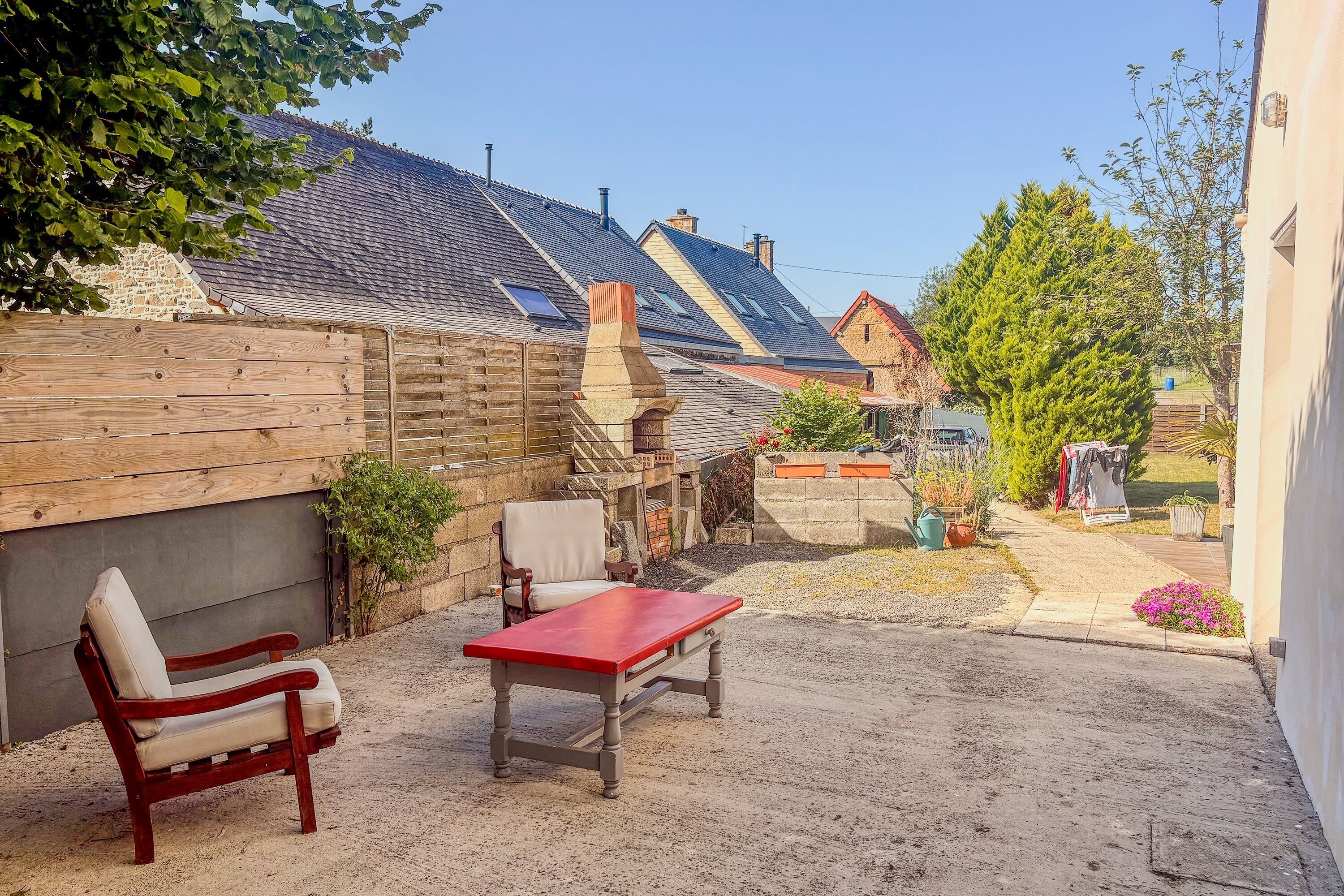 A patio with a wooden table and two chairs, a stone wall, and a house with a tiled roof in the background.