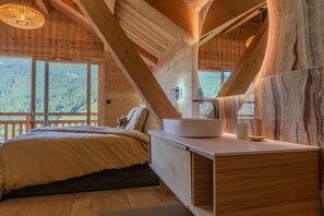 A wooden cabin bedroom with a large bed, a bedside table with a bowl, and a view of a forest through large windows. - Jag Peak - Five Bedroom House, Sleeps 12 (Saint-Chaffrey)