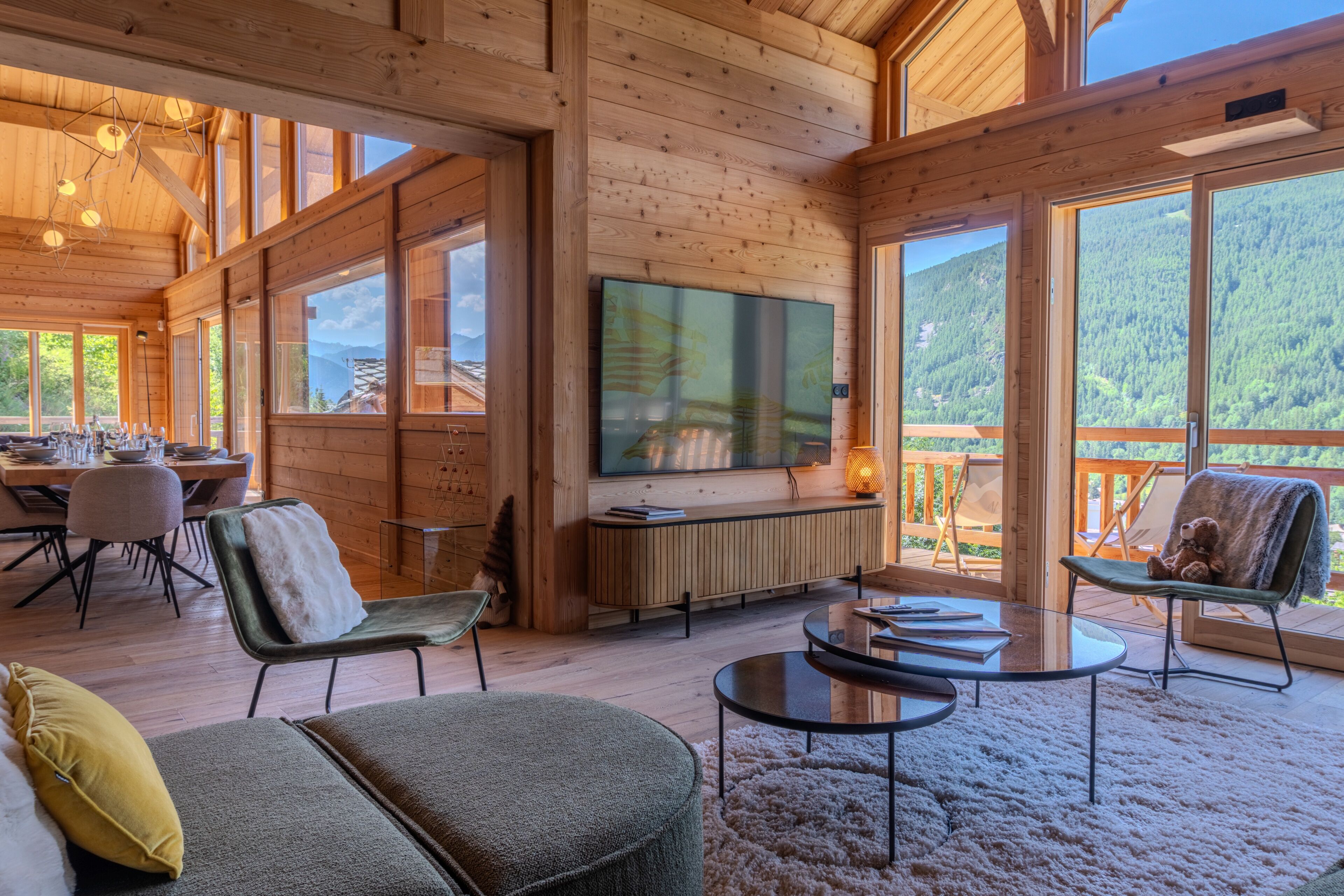 A wooden cabin interior with a TV, seating area, dining space, and large windows.