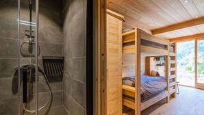 Ein modernes Badezimmer mit Duschkabine aus Glas, einem Holzbett mit Schlafnische und Schreibtisch sowie einem großen Fenster mit Blick ins Freie.