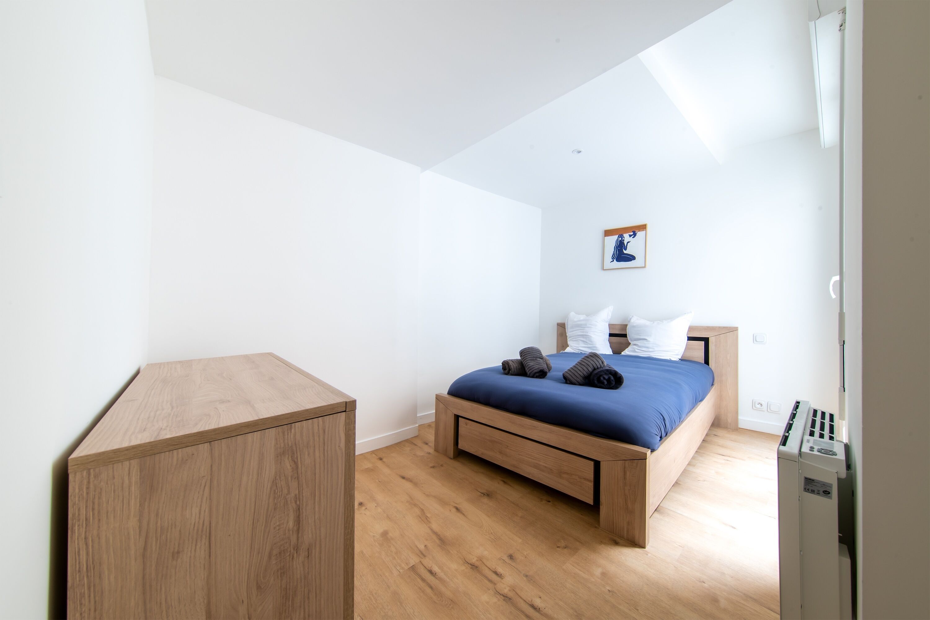 Une chambre à coucher avec un lit en bois, un petit bureau en bois et un mur blanc orné d’un tableau encadré.