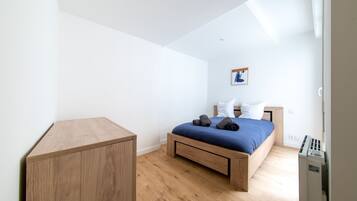 Une chambre à coucher avec un lit en bois, un petit bureau en bois et un mur blanc orné d’un tableau encadré.