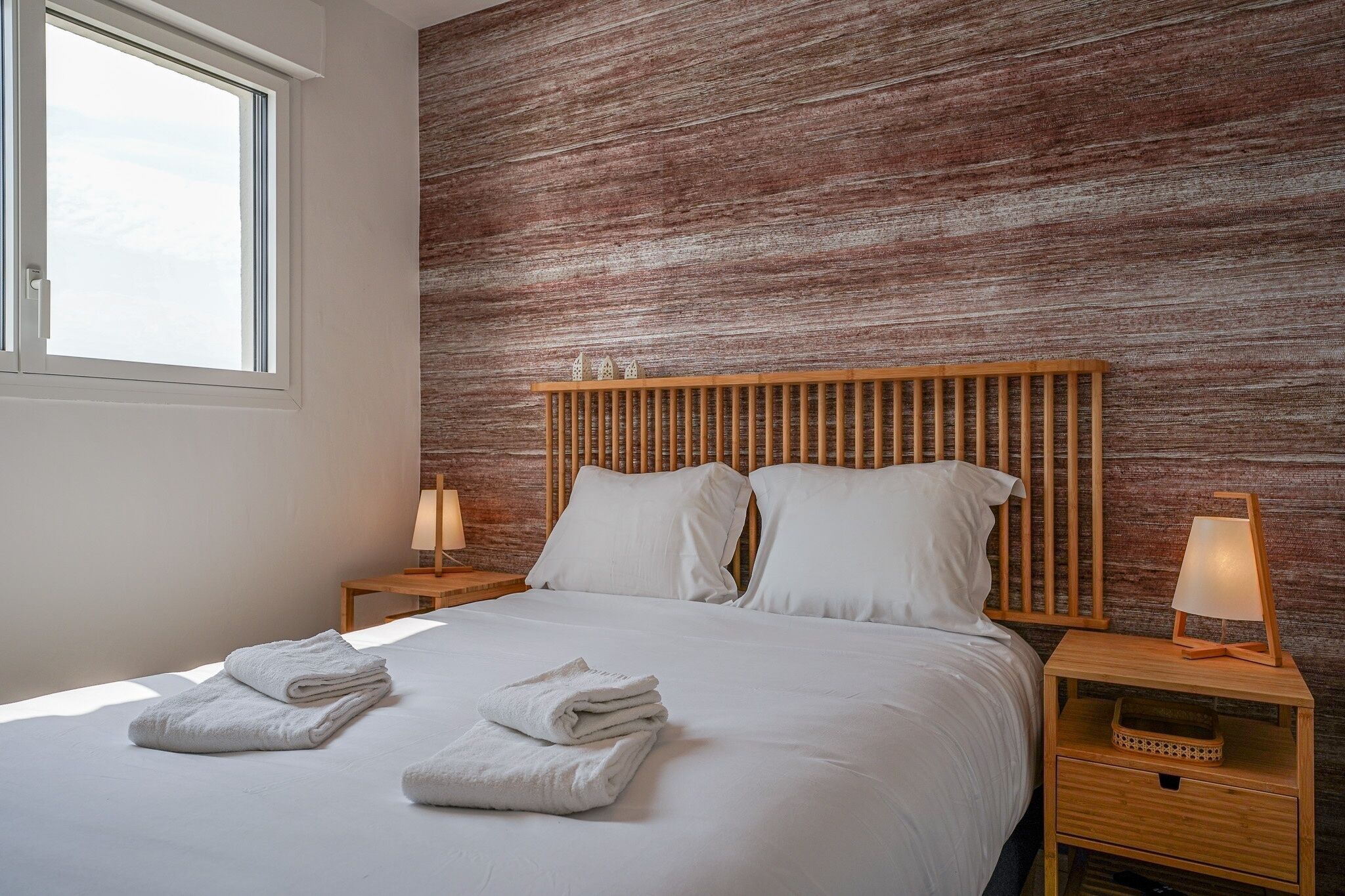 A neatly made bed with white linens and towels, a wooden headboard, two bedside tables with lamps, and a window providing natural light.