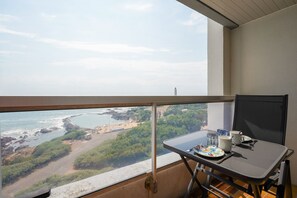 Ein Balkon mit Tisch für zwei Personen, Blick auf einen Strand mit felsiger Küste und viel Grün.