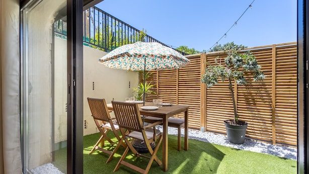 Une terrasse avec une table et des chaises en bois, un parasol à motifs et une plante en pot.